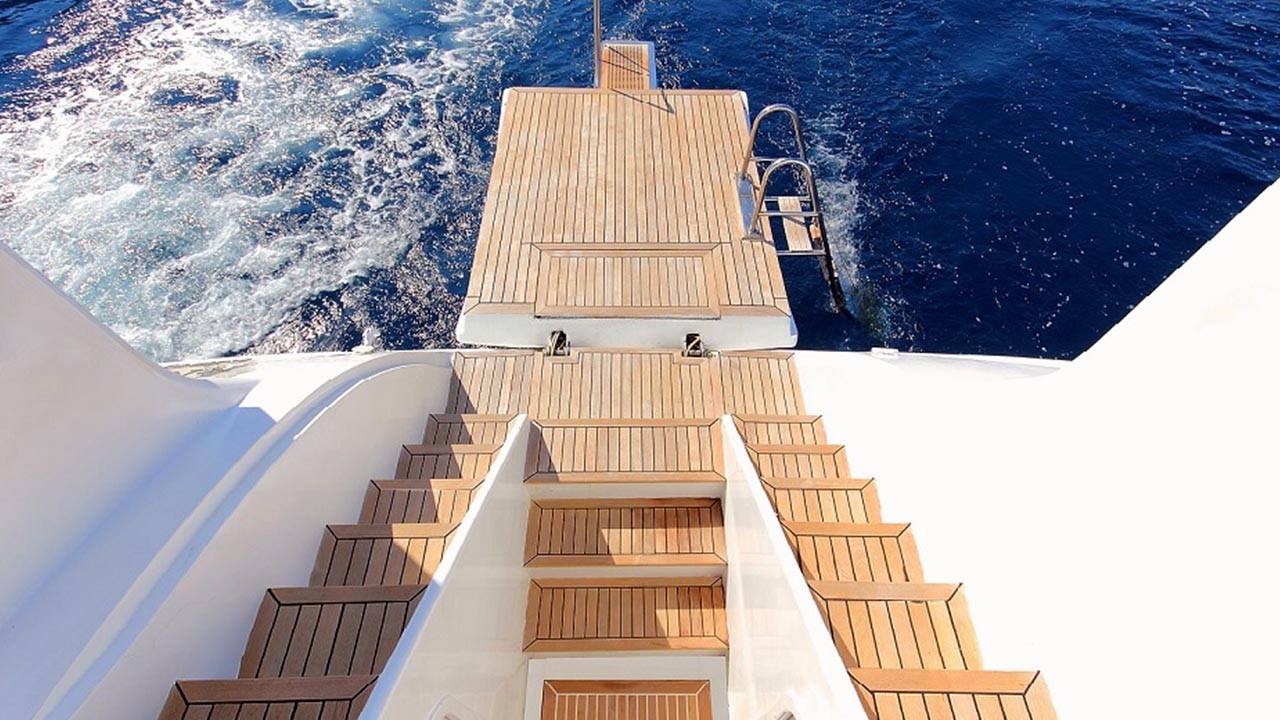 ANGELO II A view from the back of a yacht shows wooden steps leading down to a swim platform above deep blue ocean water, with a metal ladder extending into the sea. White yacht rails frame the scene.