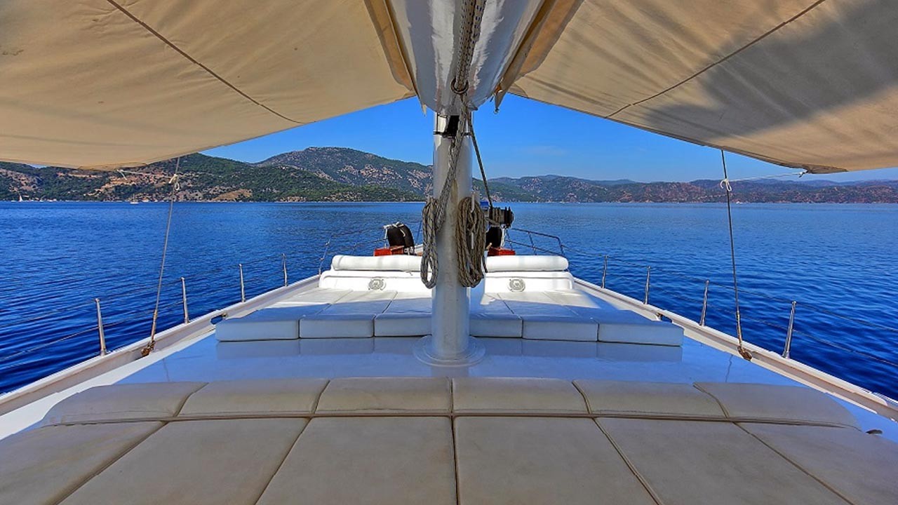 ANGELO II View from the deck of a sailboat with tan cushions and large sails, looking out over calm blue water toward distant green hills under a clear blue sky.