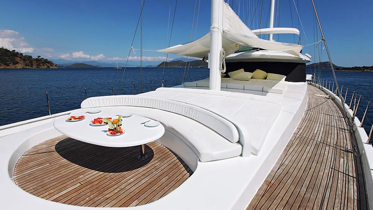 ANGELO II Luxury yacht deck with white curved seating, sunbeds, and a round table set with fruit and drinks, overlooking calm blue water and distant green hills under a clear sky.
