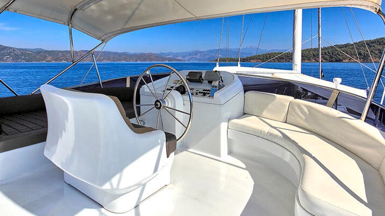 ANGELO II The helm of a yacht featuring a large white captain’s chair, a steering wheel, an instrument panel, and a curved cream-colored cushioned seating area, with blue sea and distant hills in the background.
