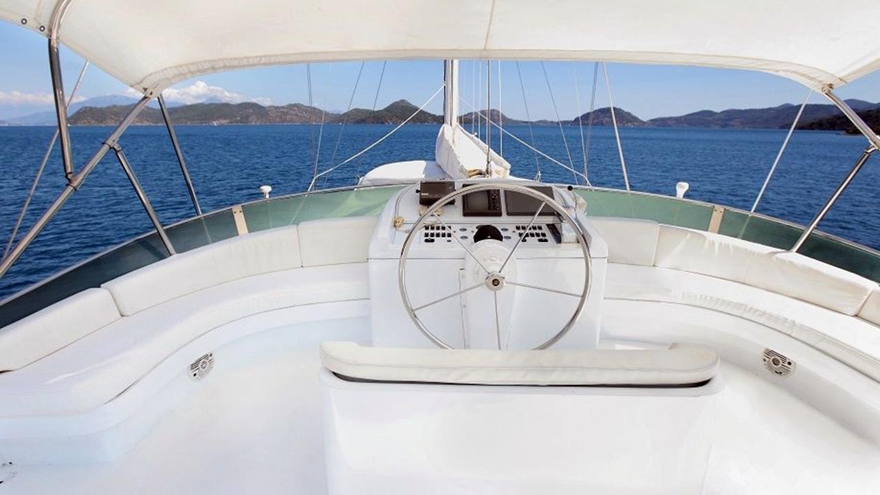 ANGELO II A view from the helm of a white yacht, showing a large steering wheel, cushioned seating, and an open canopy, with calm blue sea and islands visible in the distance under a clear sky.