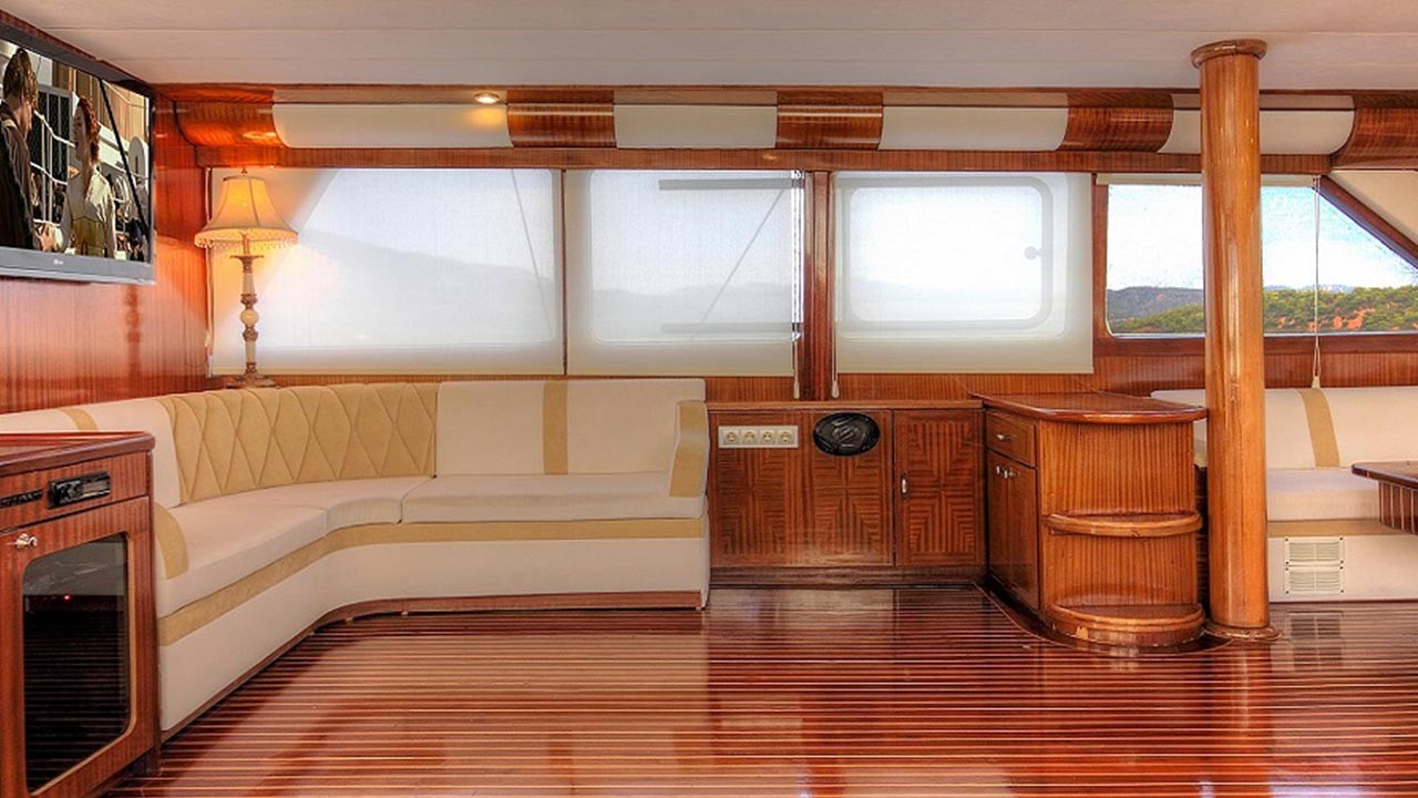ANGELO II A spacious yacht interior with polished wooden floors, cream and beige upholstered seating, large windows, wood cabinetry, a standing lamp, and a wall-mounted TV. Natural light fills the room.