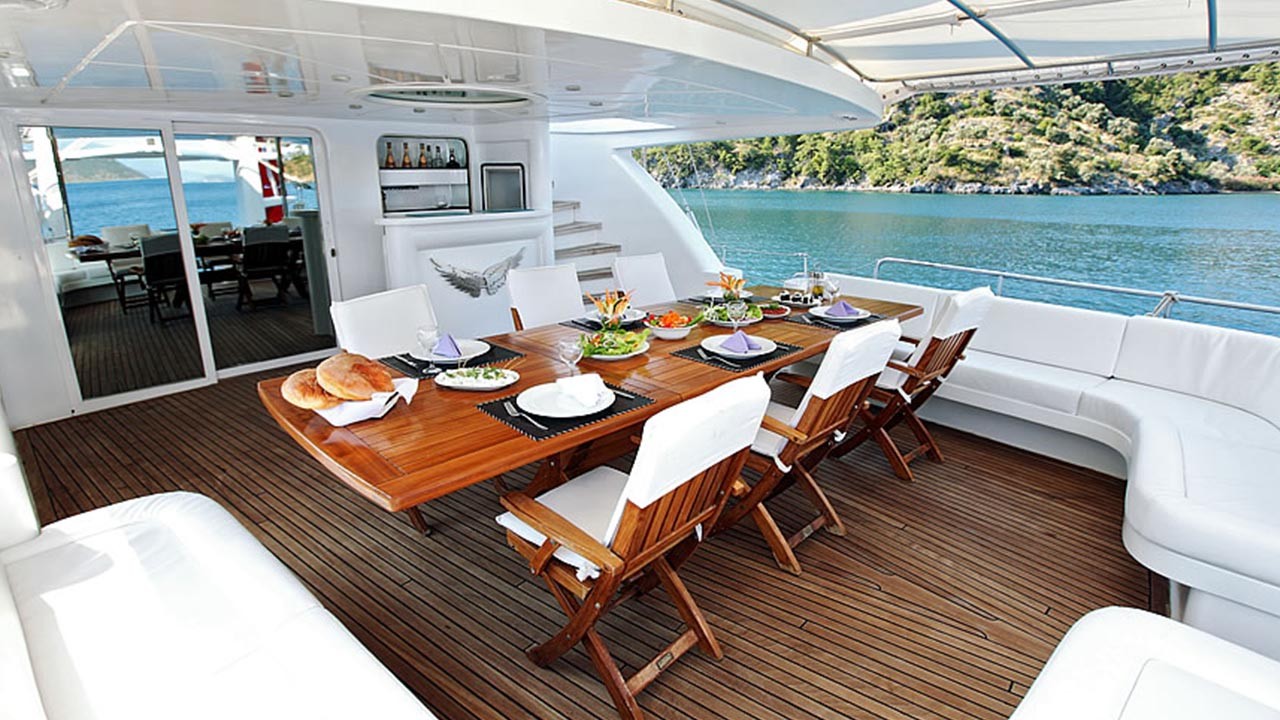 ANGELO II A luxurious yacht dining area with a wooden table set for a meal, surrounded by white cushioned chairs, overlooking calm blue water and a green, forested coastline.