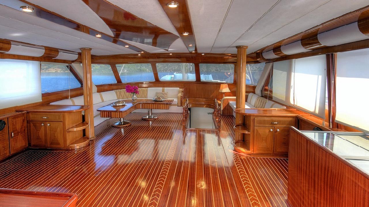 ANGELO II Spacious yacht interior with polished wooden floors, large windows, and white sofas around a coffee table. Natural light fills the room, highlighting the elegant and luxurious design.