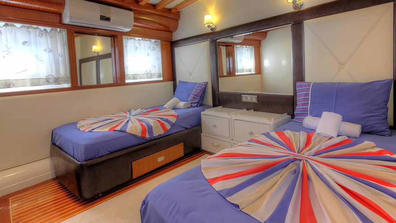 ANGELO II A cozy cabin with two single beds, each with blue bedding and colorful round covers. Rolled towels are placed on one bed. There are large mirrors, a white dresser, wood accents, and windows with patterned curtains.