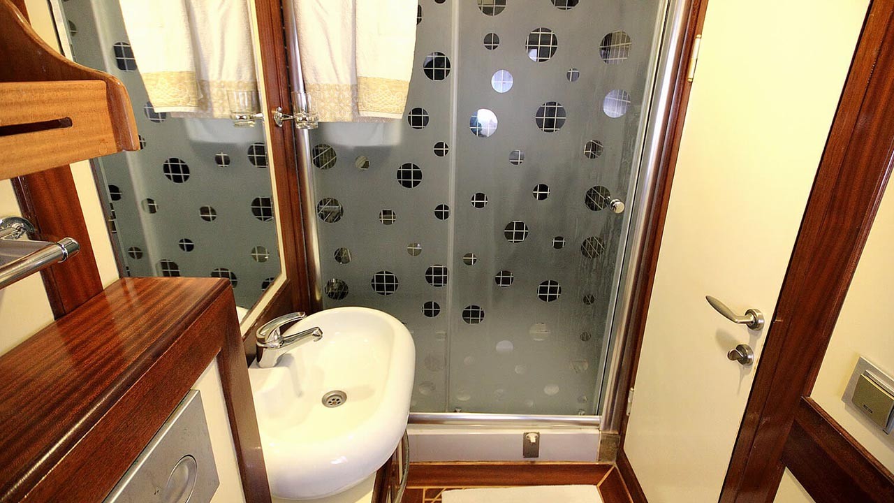 ANGELO II Small bathroom with a white sink, a wall-mounted mirror, wooden details, a towel on a rack, and a shower area enclosed by frosted glass with circular patterns. A white door is partly visible on the right.