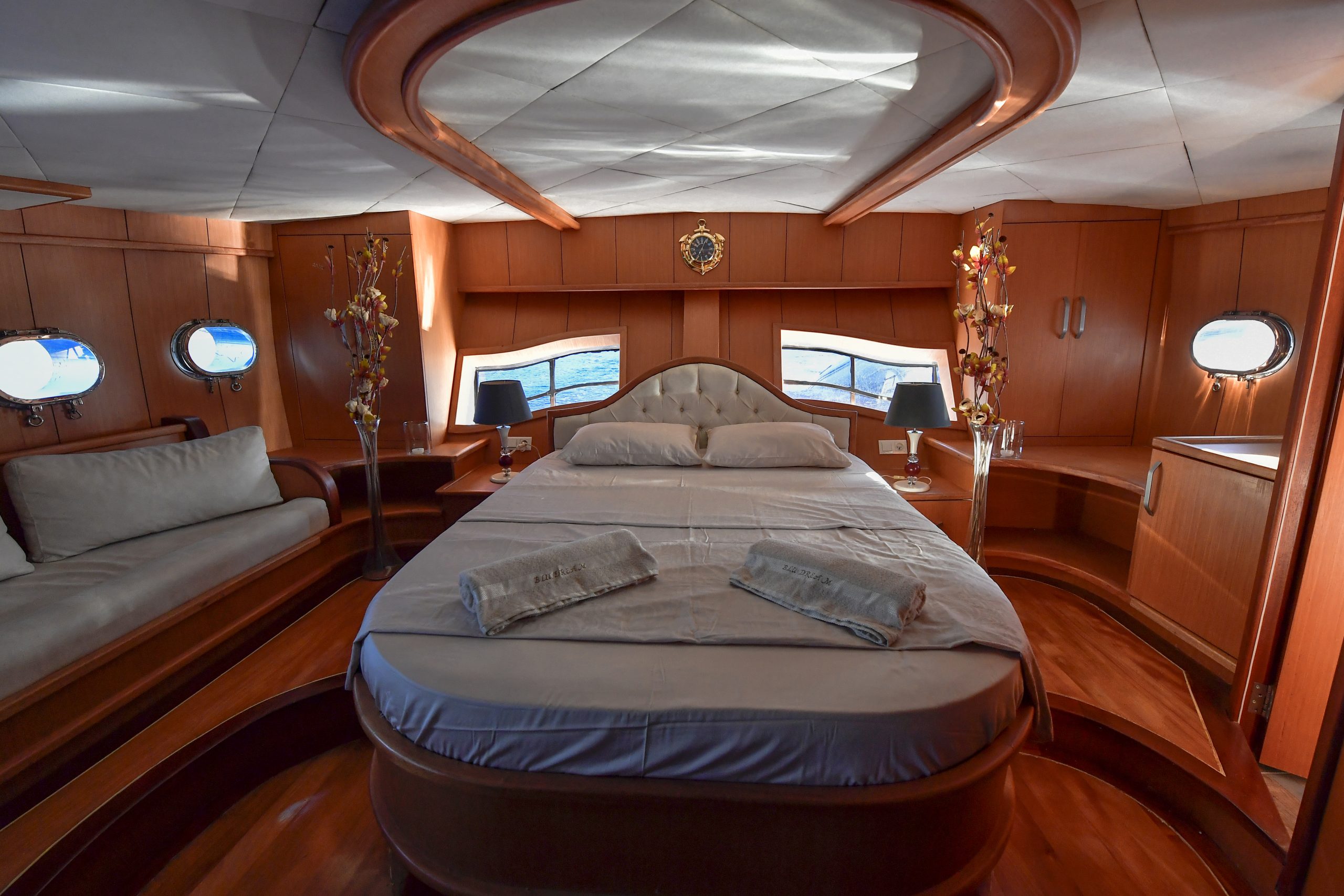 Luxurious bedroom with a large bed, beige headboard, sofa, wood-paneled walls, built-in cabinets, small round windows, and soft lighting, creating an elegant and cozy atmosphere.