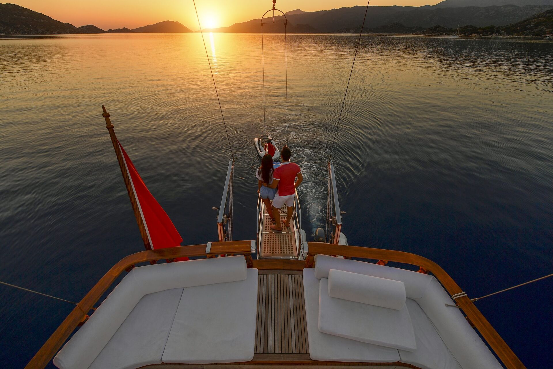 A couple stands at the back of a yacht, holding hands and looking at the sunset over calm water and distant mountains, with a red flag visible on the boat.