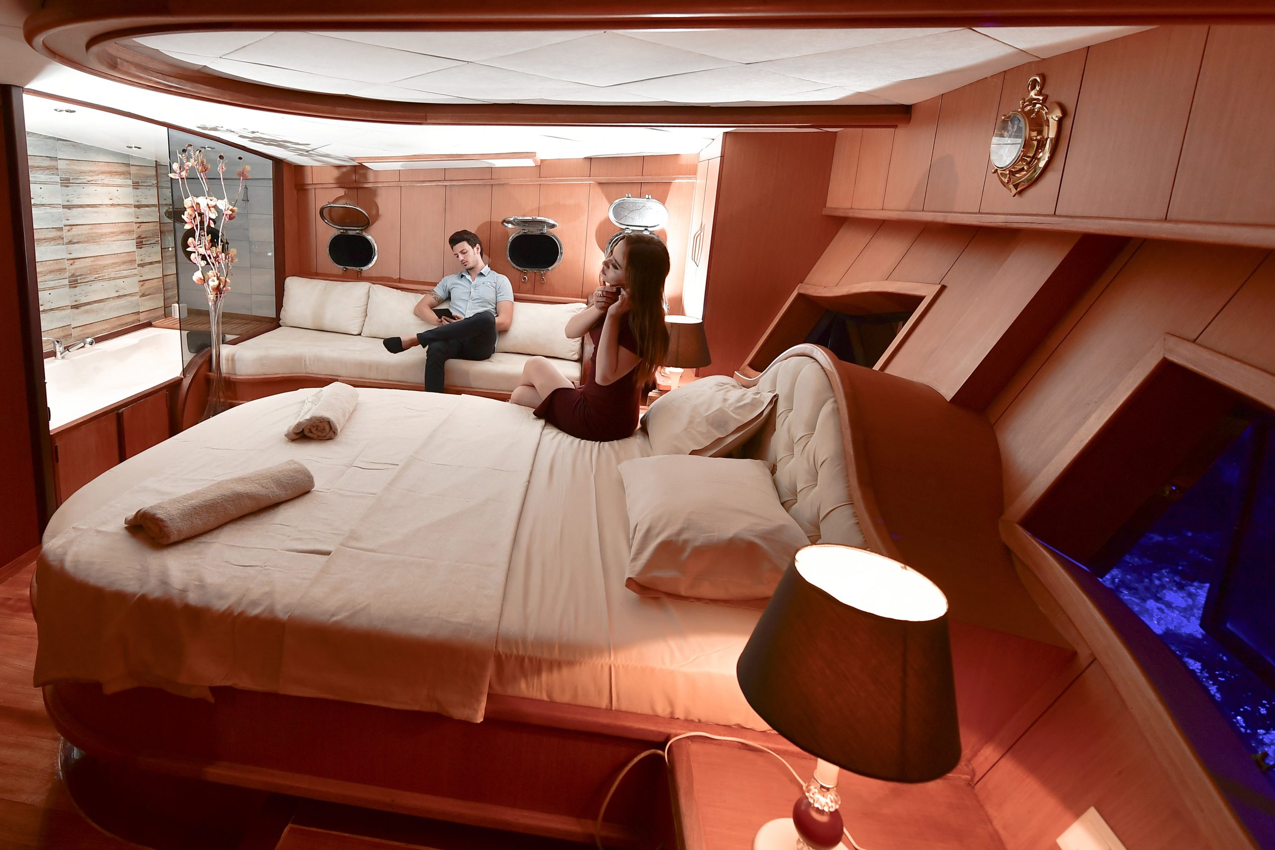 A woman sits on a large bed in a luxurious yacht bedroom while a man lounges on a sofa near a window; the room features warm wooden decor, soft lighting, and a view of water through a porthole.