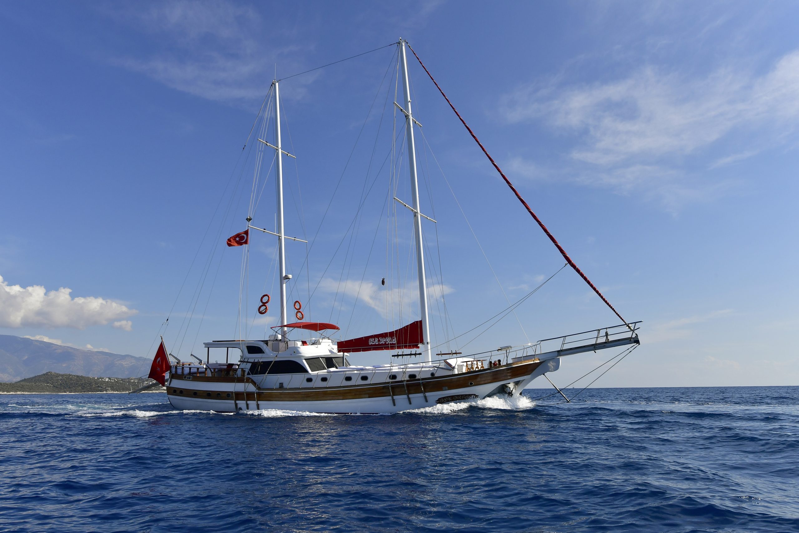 A large white sailing yacht with two tall masts and Turkish flags cruises on deep blue water under a clear sky, with distant mountains visible on the left.