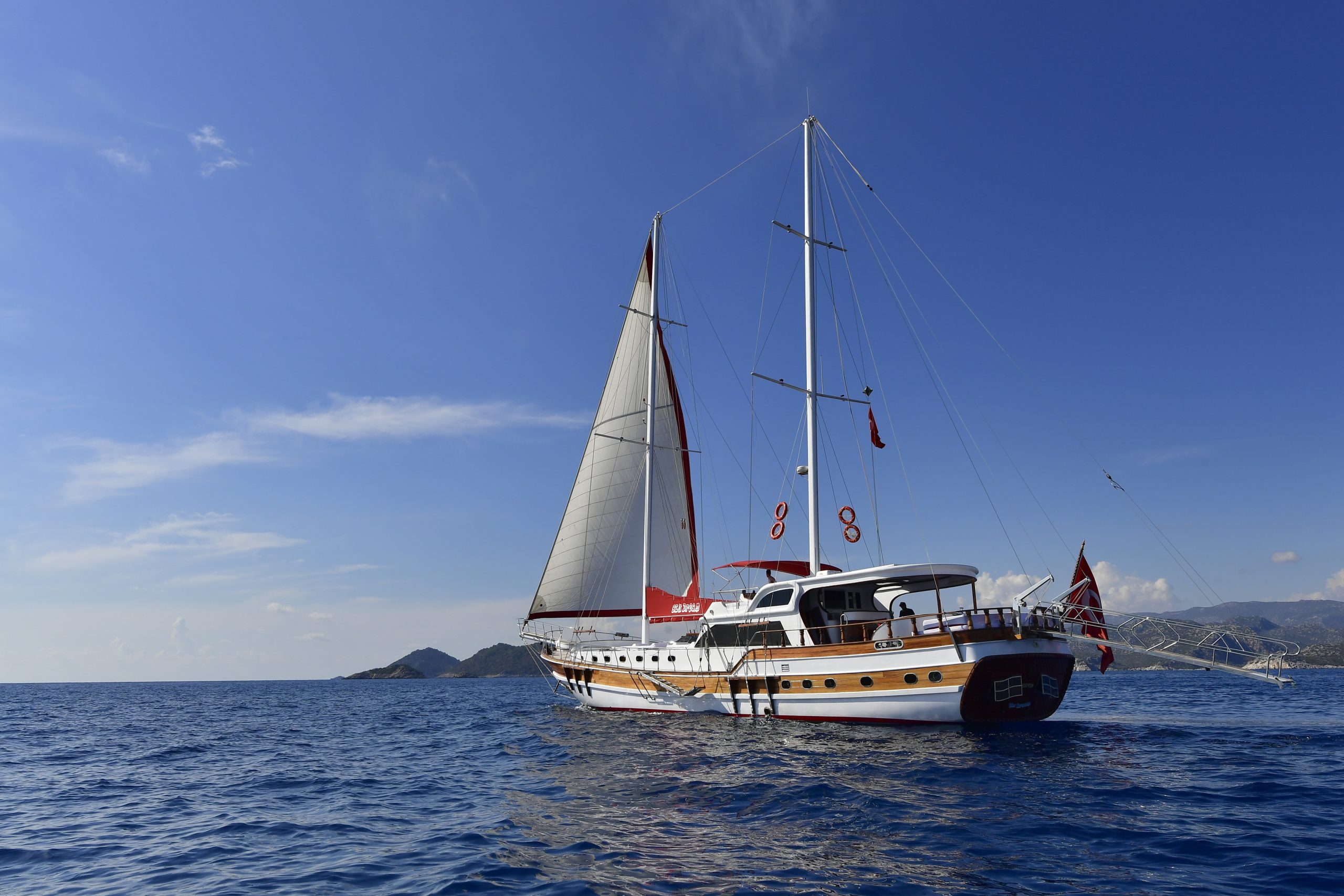 A white and wooden sailboat with raised sails floats on calm blue water under a clear sky, with distant hills visible on the horizon.