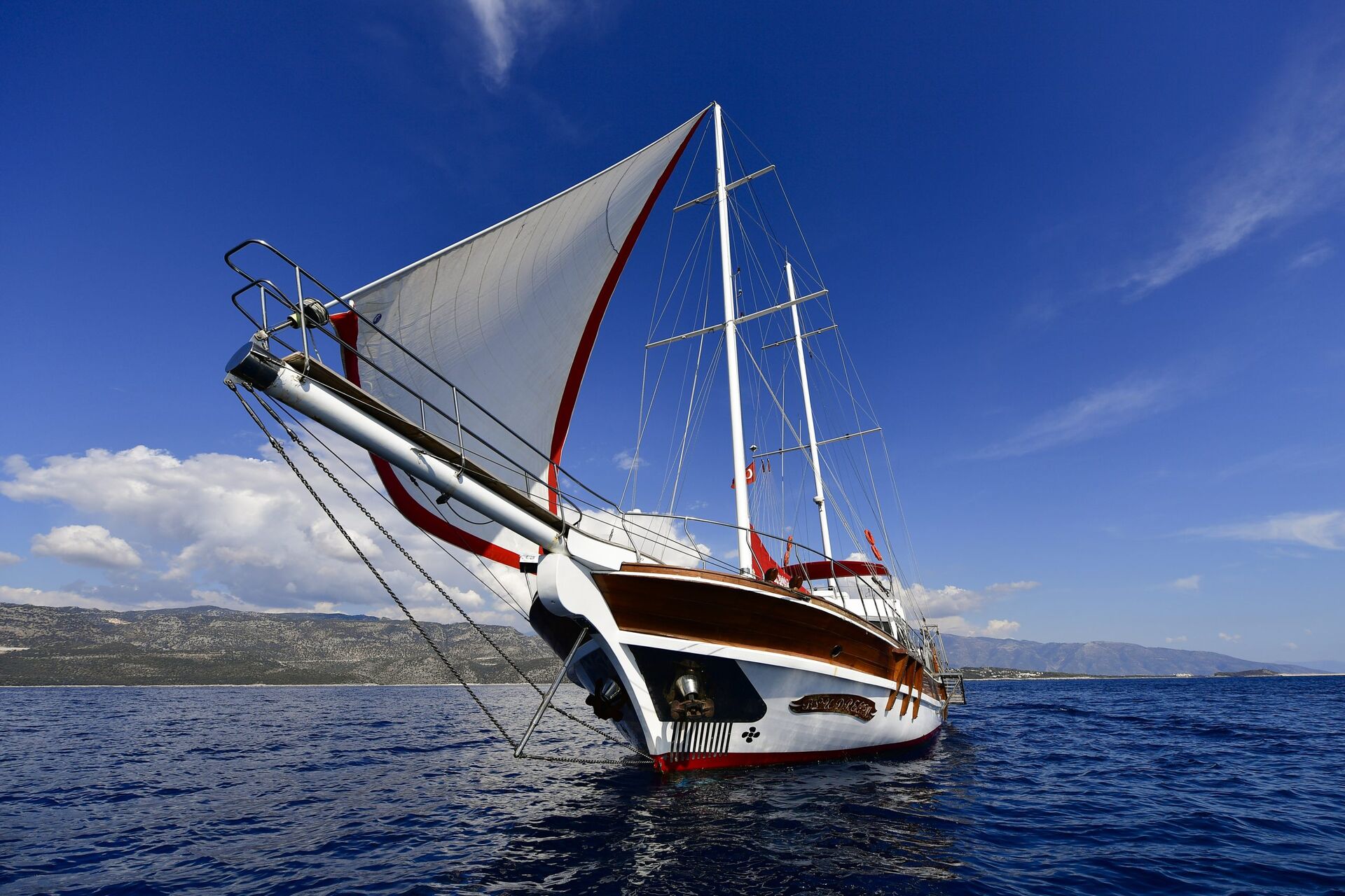 A large wooden sailboat with white and red sails is anchored on calm blue water under a clear sky, with distant mountains and clouds in the background.