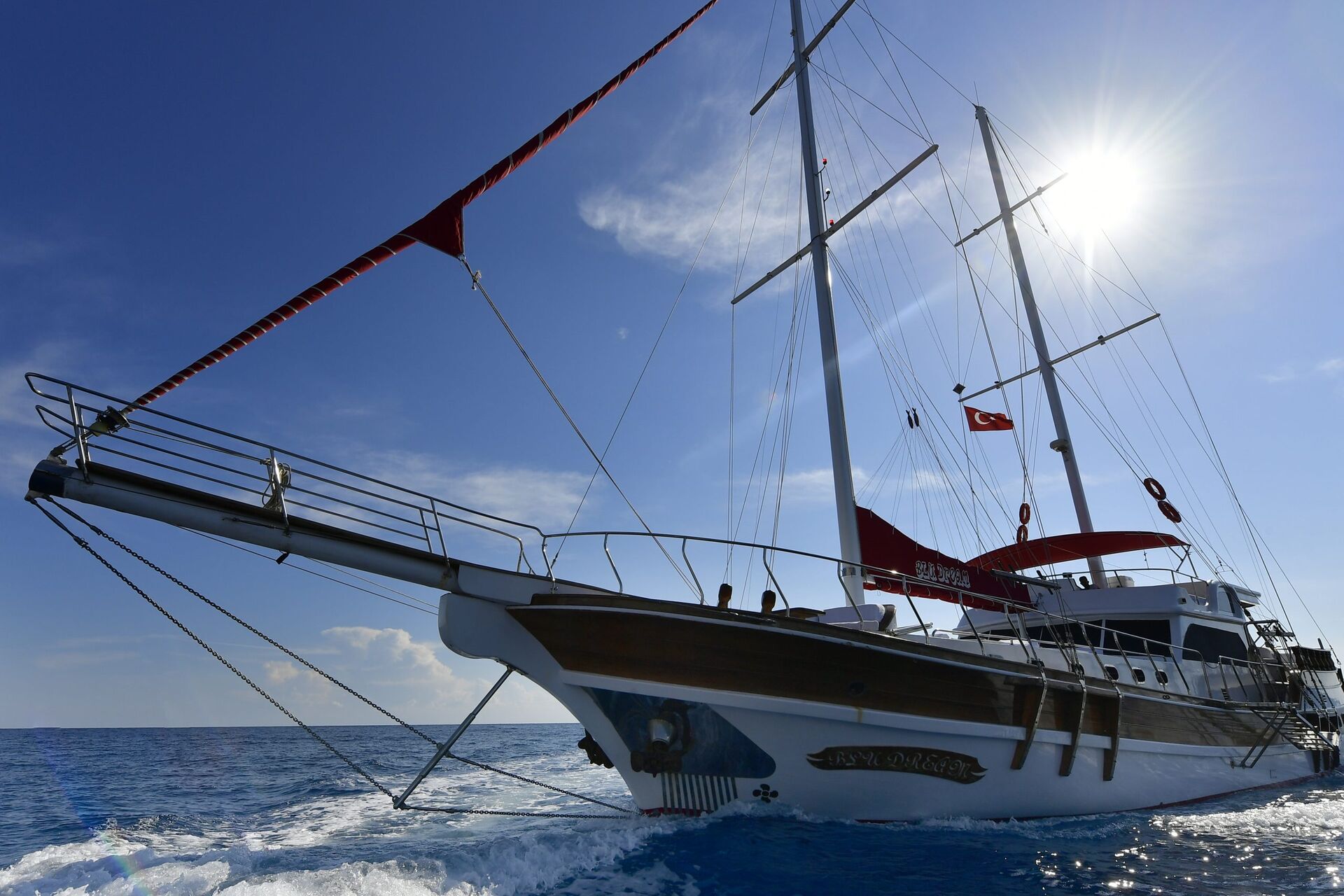 A large white and wooden sailboat with Turkish flags sails on blue ocean water under a bright sun and clear sky. The boat creates waves as it moves forward.