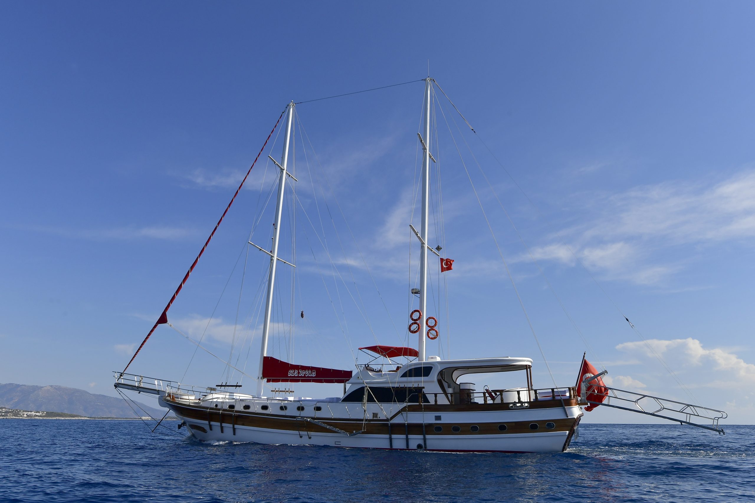 A white and brown sailing yacht with two tall masts floats on calm blue water under a clear sky, with a Turkish flag flying at the stern. Land is visible in the background on the left.