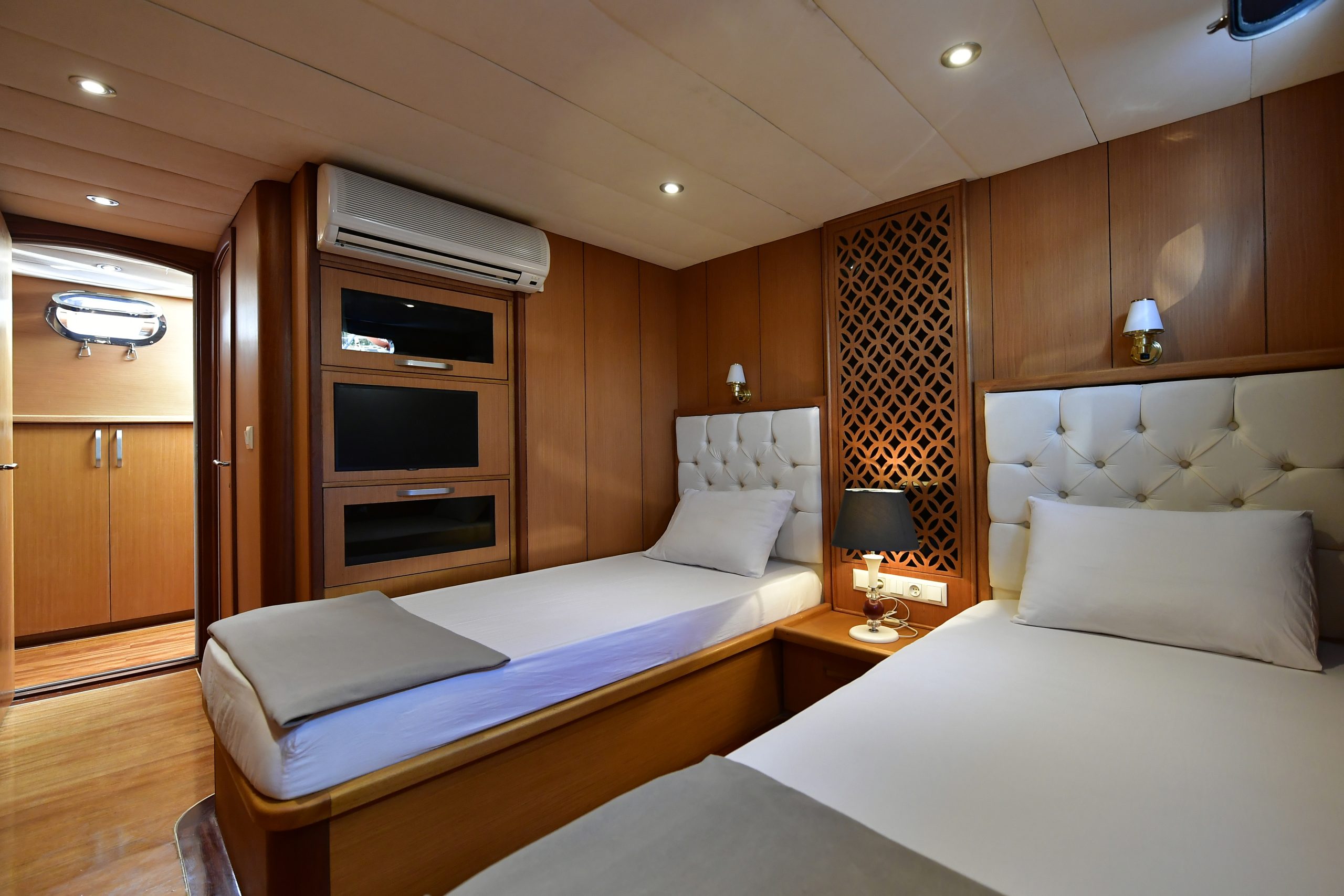 A cozy yacht cabin with two single beds, white bedding, a wooden nightstand with a lamp, wooden walls, and built-in shelves. A small window and soft ceiling lights brighten the room.