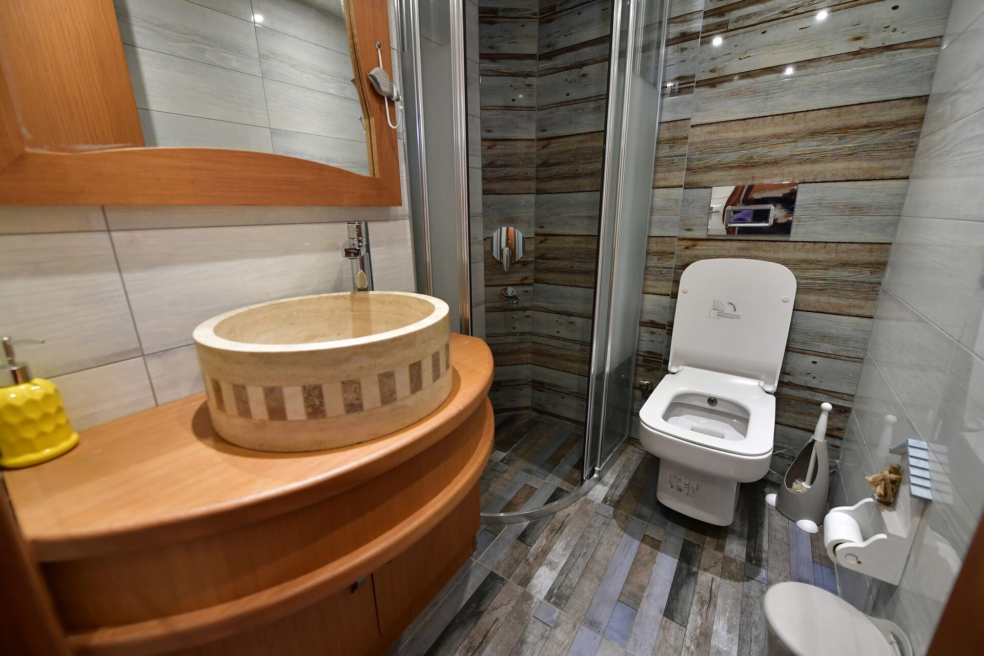A modern bathroom with wood-patterned walls, a round stone sink on a wooden counter, a toilet, a glass-enclosed shower, and toiletries including soap and toilet paper.