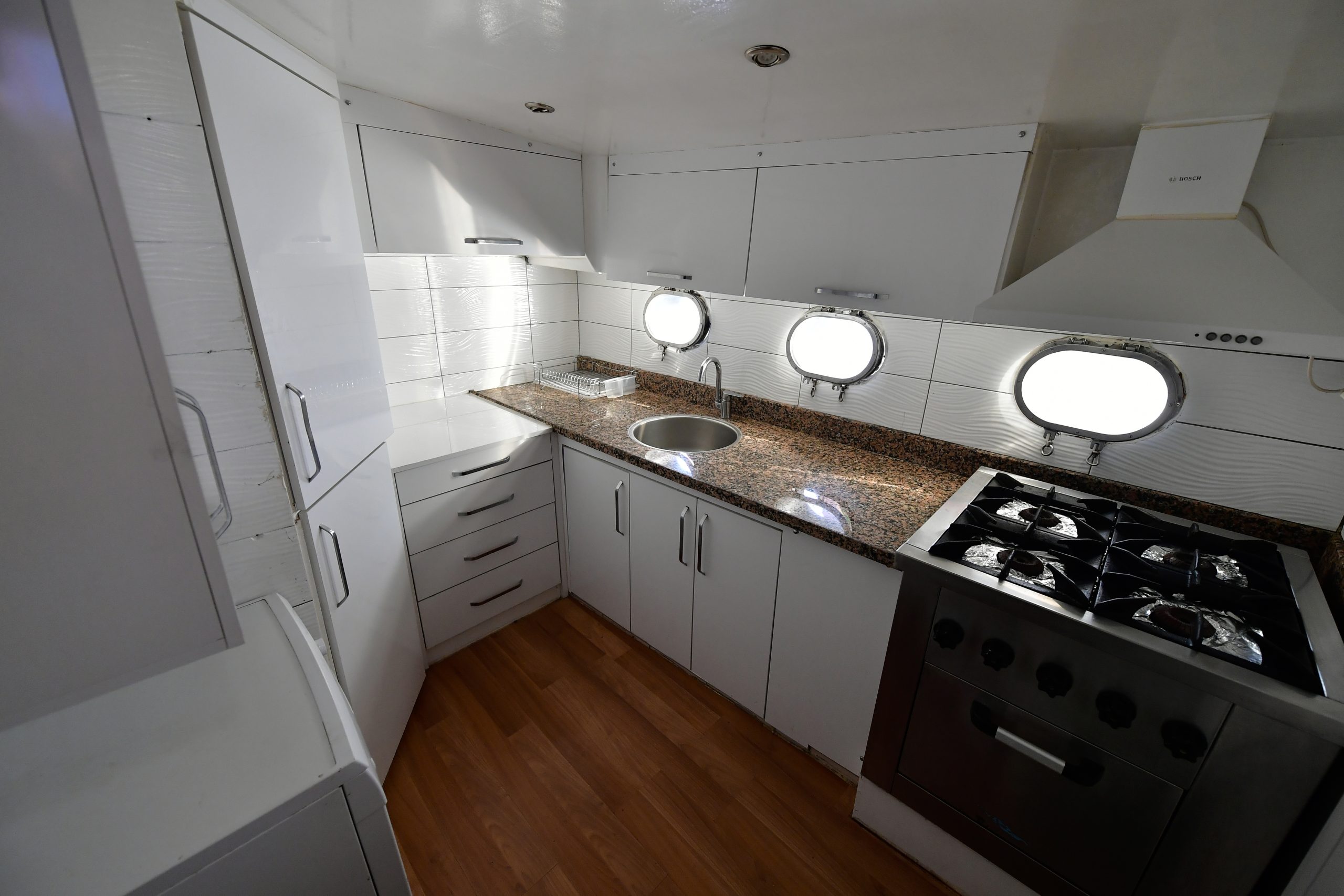 Modern kitchen with white cabinets, granite countertops, a gas stove, sink, and three round windows. Wood flooring contrasts with the bright, clean design.