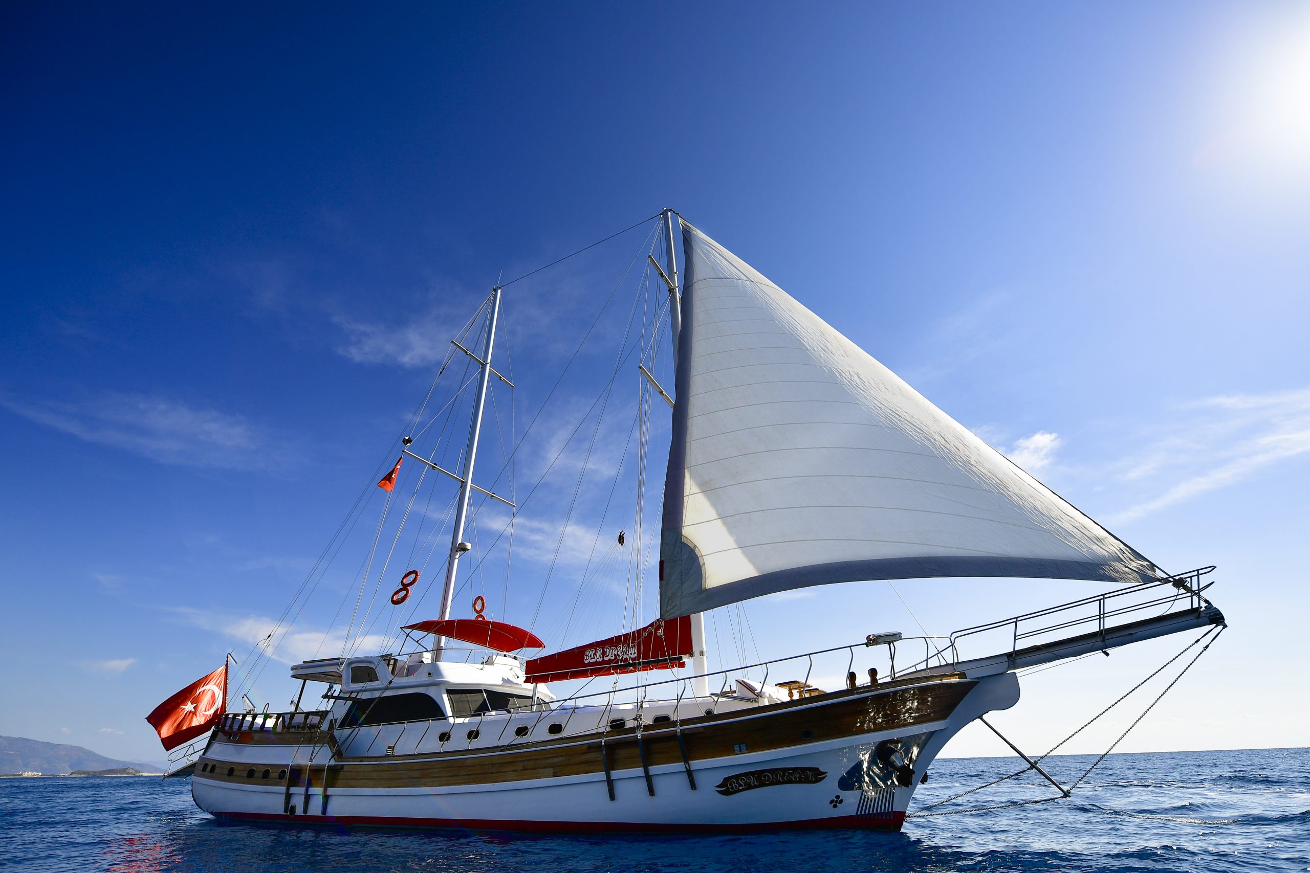 A wooden sailboat with a large white sail glides on clear blue water under a bright sky. Red flags are visible on the boat, and distant land can be seen on the horizon.