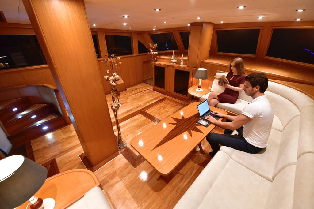 A man works on a laptop and a woman reads a magazine in a stylish yacht lounge with wooden floors, modern furnishings, and large windows.