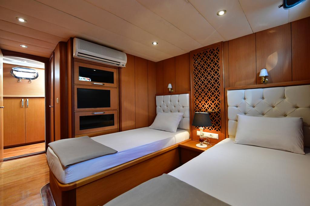 A cozy cabin with two single beds, padded white headboards, wood-paneled walls, built-in shelves, a lamp on a bedside table, and an air conditioner above. A small window lets in natural light.