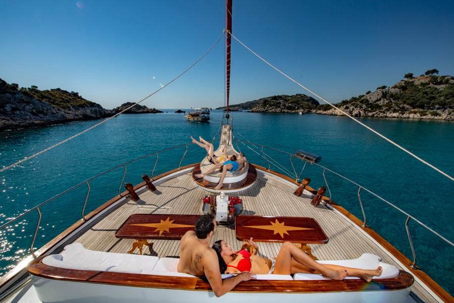 People relax on the deck of a yacht anchored in clear turquoise water, surrounded by rocky green islands under a bright blue sky. Some are sunbathing on loungers, enjoying the peaceful, scenic setting.