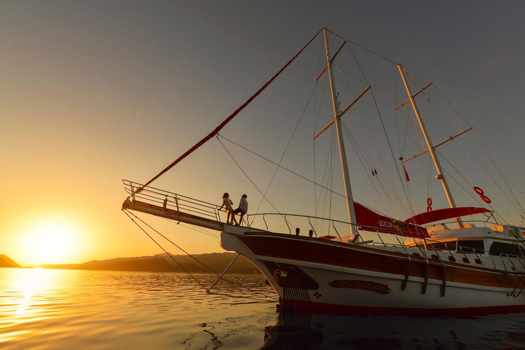 A couple stands at the bow of a large sailboat during sunset, with the sun low over calm water and distant hills, creating a warm, golden glow over the scene.