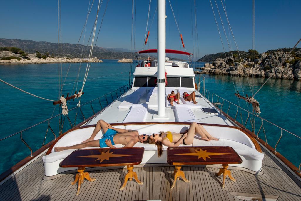 Four people relax on the deck of a yacht under clear blue skies, surrounded by calm turquoise water and rocky islands. Two people are sunbathing at the front, while two more lounge on sunbeds farther back.