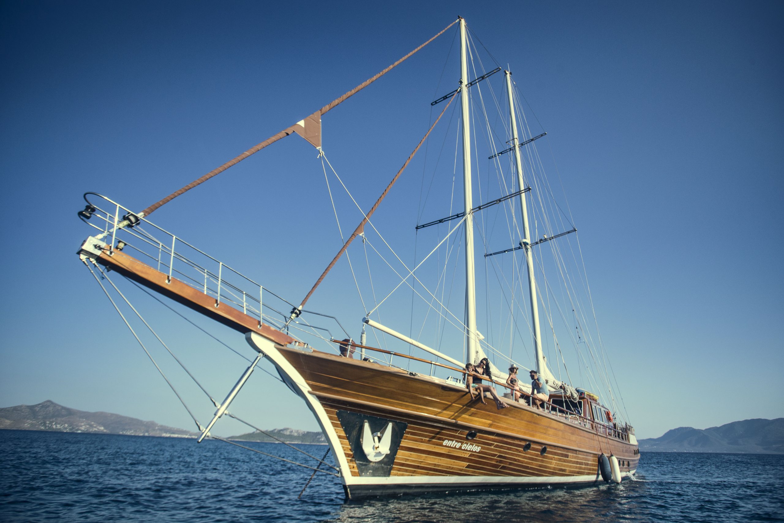 A large wooden sailboat with tall masts sails on calm blue water under a clear sky, with distant hills visible in the background. Several people are on deck enjoying the sunny day.