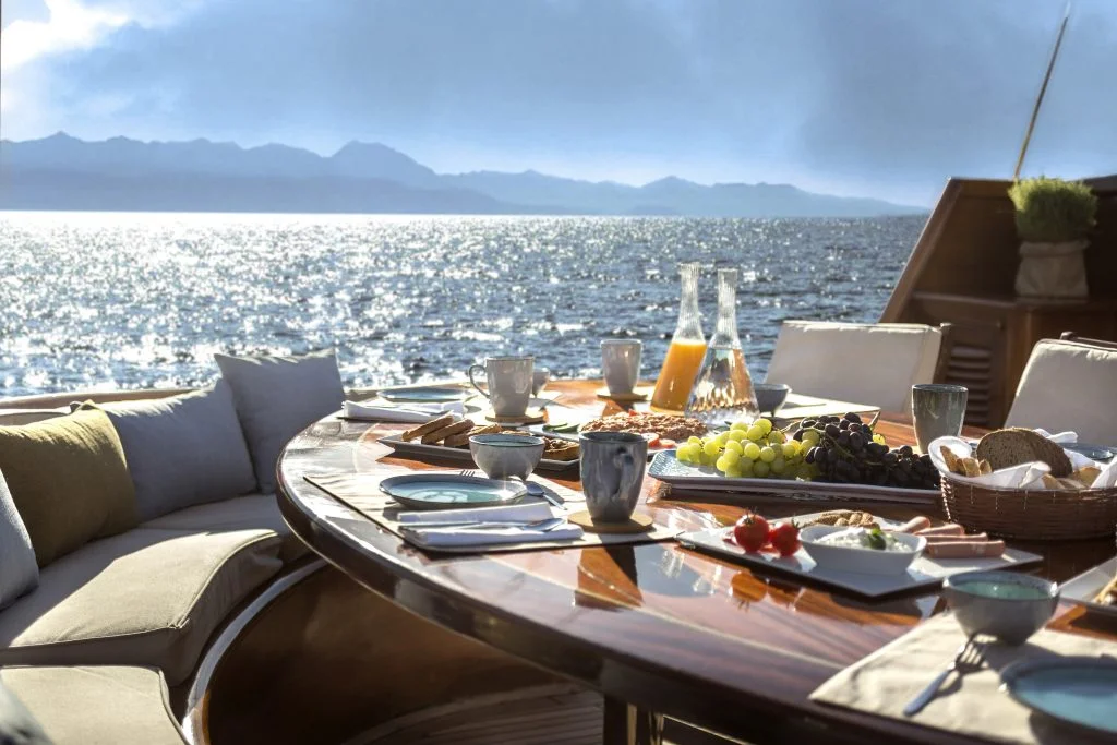 A sunlit outdoor table on a yacht, set for breakfast with juice, fruit, cheese, bread, and cups, overlooks a sparkling sea and mountains in the distance. Cushioned seating surrounds the table.