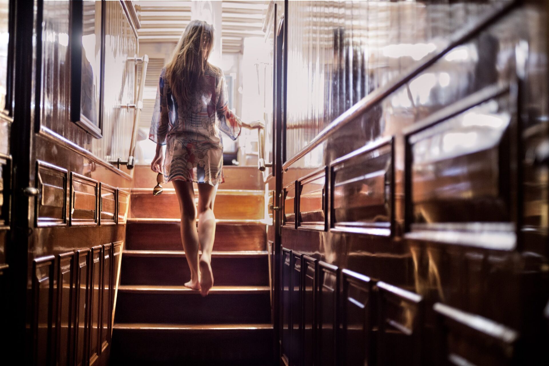 A woman in a light dress walks barefoot up wooden stairs inside a warmly lit, polished hallway, heading toward sunlight streaming in from above.