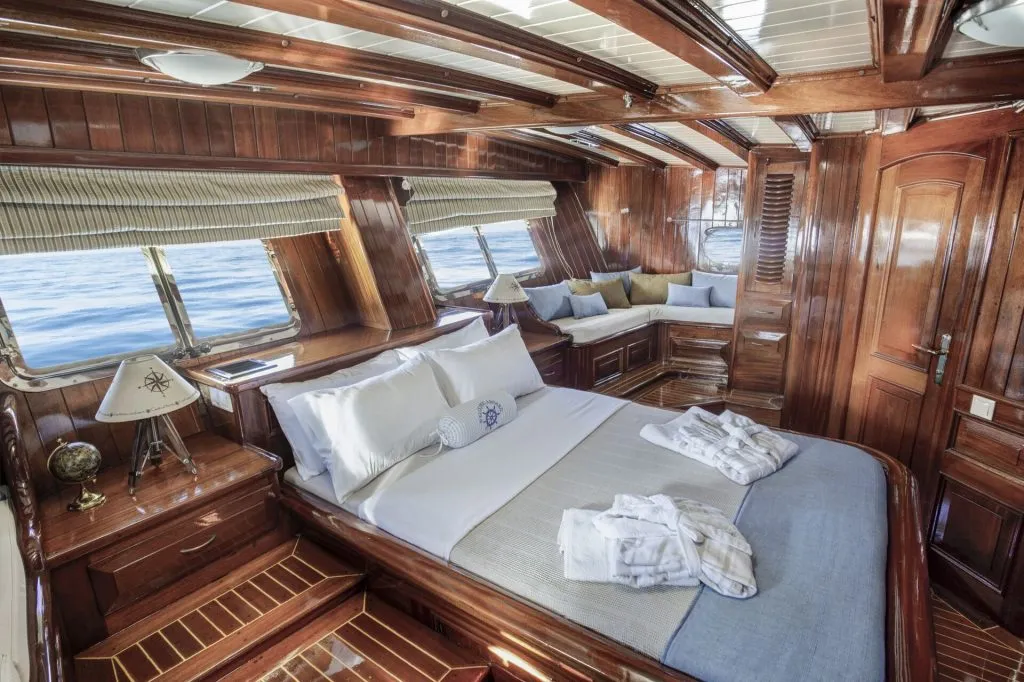 Luxurious yacht cabin with polished wood finishes, a made-up bed with white linens, two lamps, windows showing the sea, and a cozy lounge seating area by the windows.