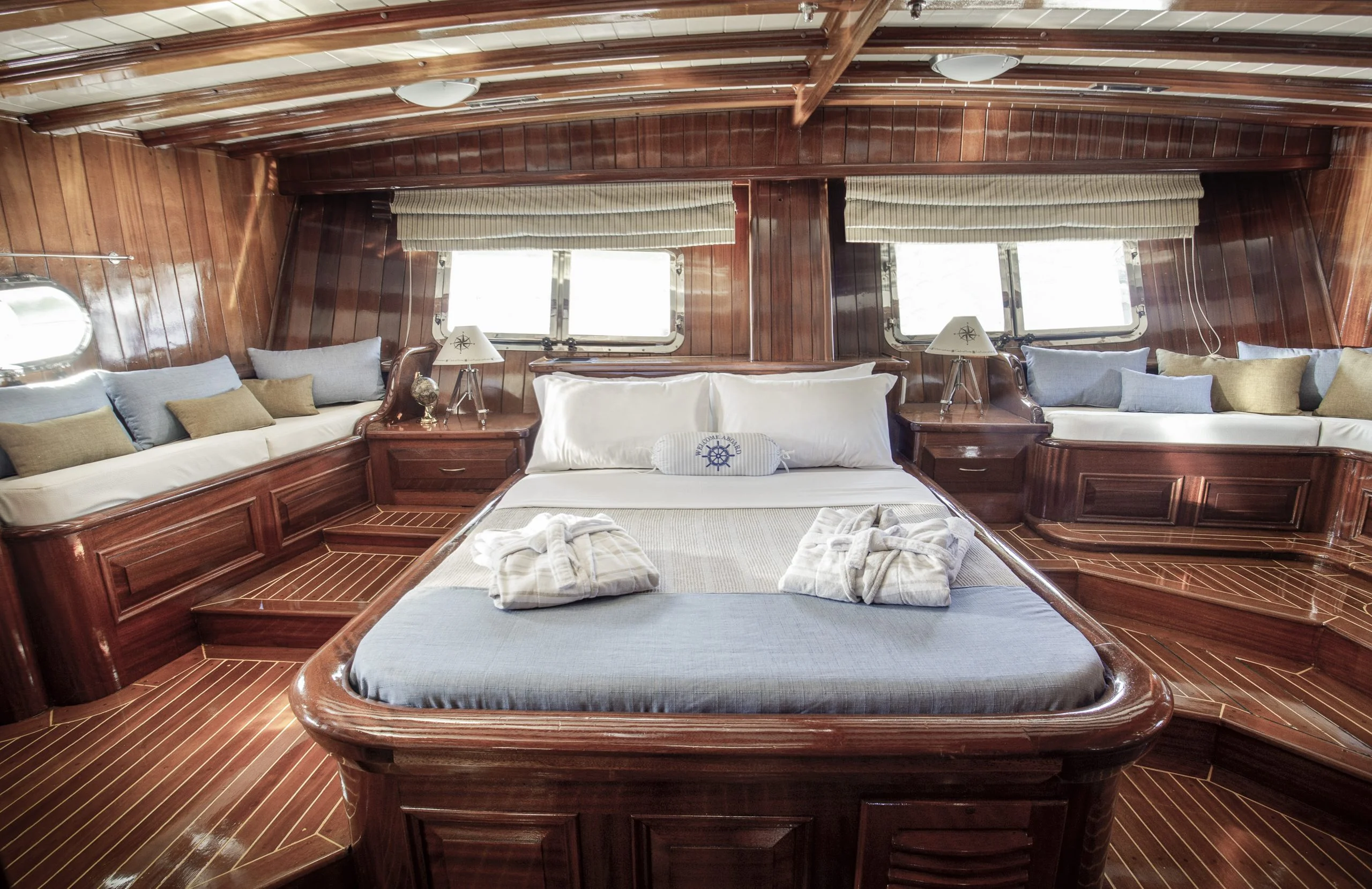 Wood-paneled yacht cabin with a large bed in the center, neatly arranged towels on top, windows behind the bed, cushioned benches on both sides, and elegant nautical decor throughout.