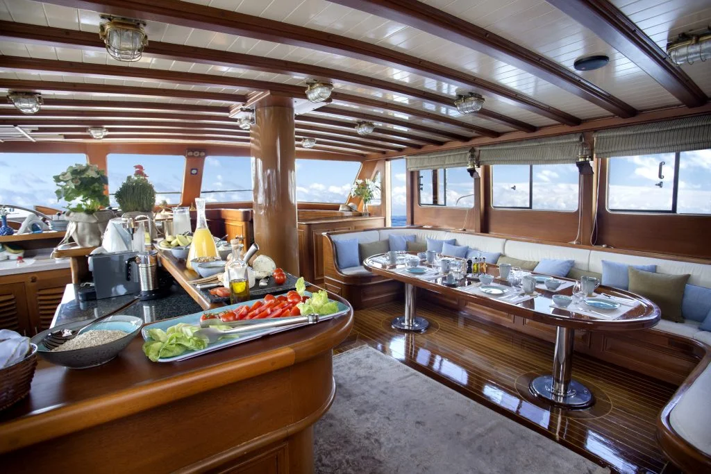 Luxurious yacht interior with polished wooden floors and ceiling, a breakfast spread on the counter, a dining table set for a meal, and large windows showing a bright, sunny sky and ocean views.