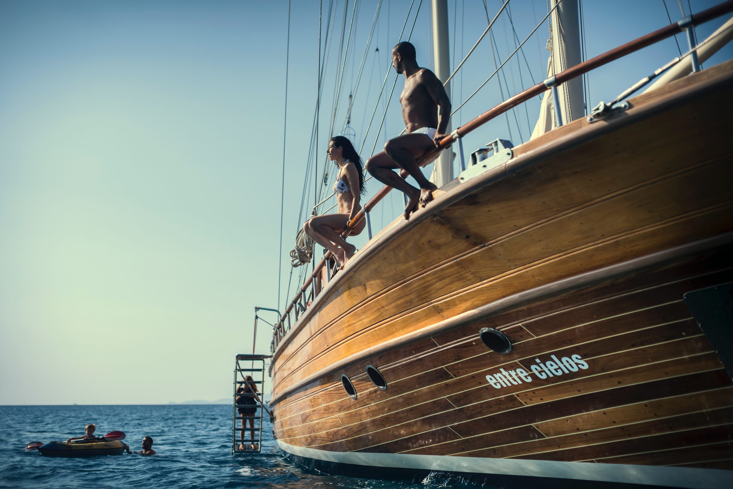 Two people prepare to jump off the side of a wooden sailboat named “Entre Cielos” into the sea, while others swim and a small inflatable boat floats nearby under a clear sky.