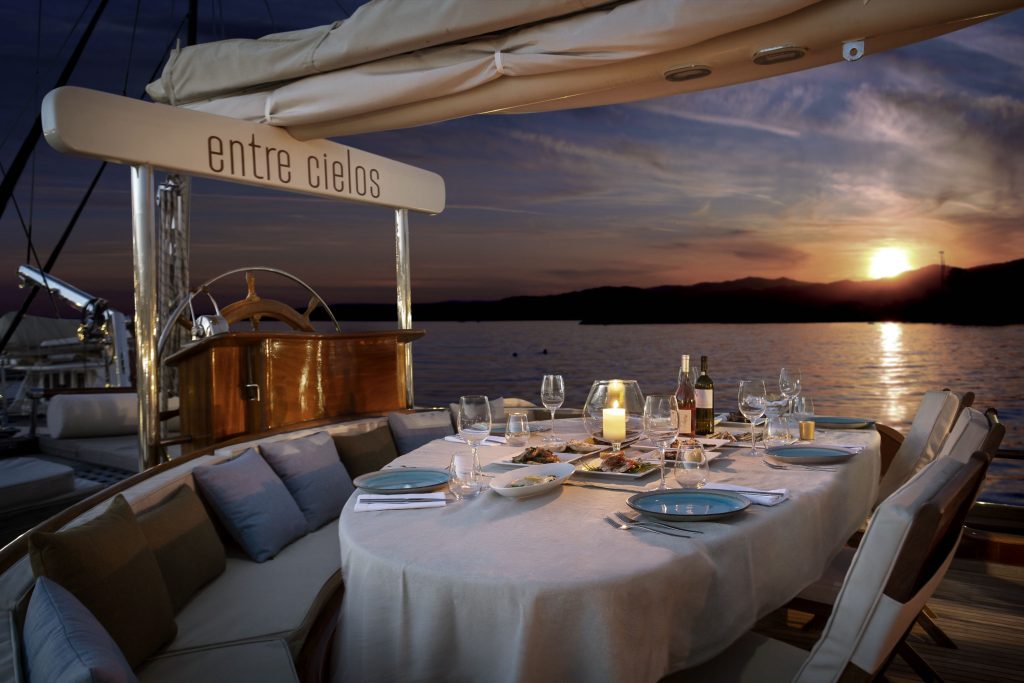 A round table set for dinner with plates, glasses, wine, and food on a yacht at sunset, with calm water and mountains in the background. The yachts sign reads entre cielos.