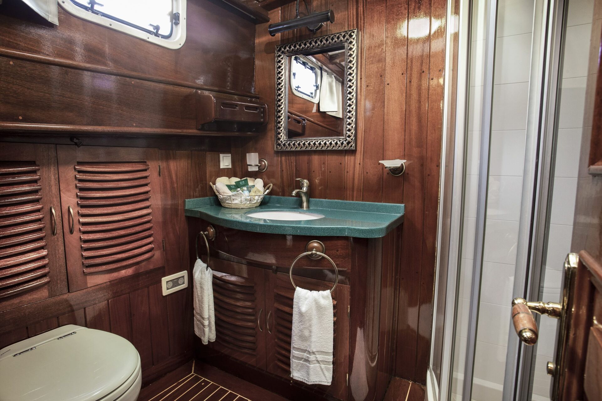 A small bathroom with rich wooden walls and cabinets, a green countertop with a sink, mirror above, white towels hanging, a window, and a glass shower enclosure visible on the right.