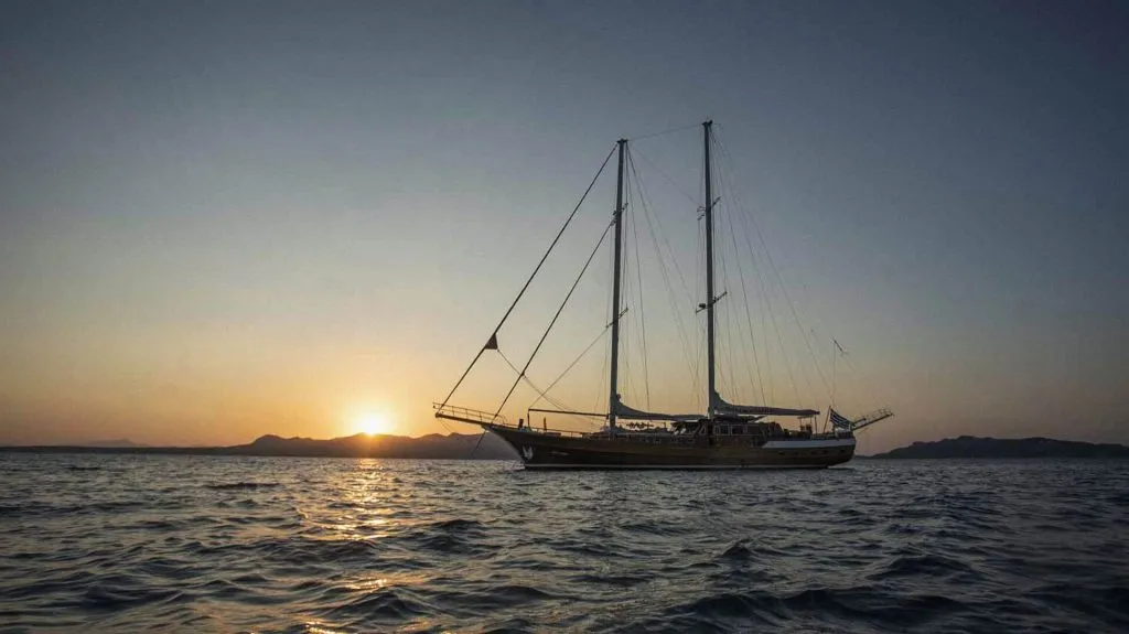 A large sailboat is anchored on calm water at sunset, with the sun setting behind distant mountains and the sky fading from orange to blue.