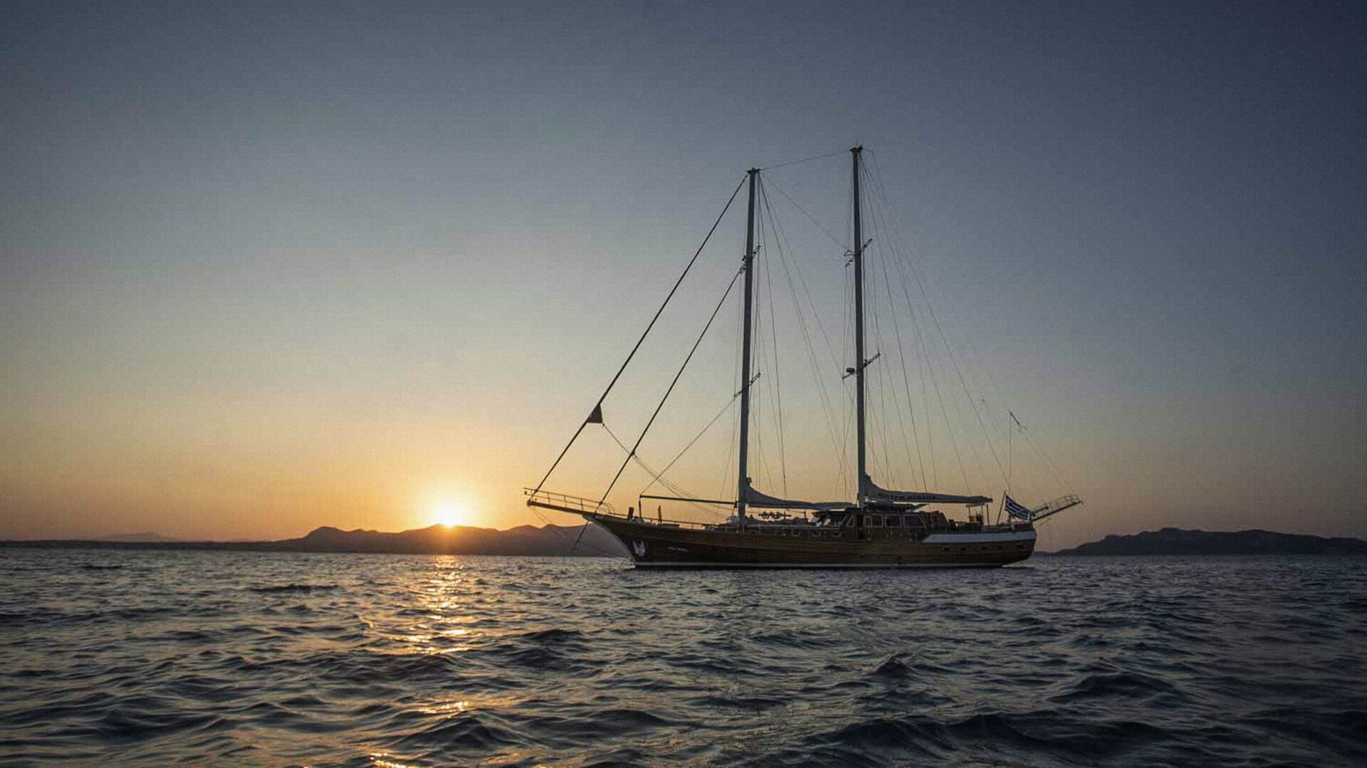 A large sailboat is anchored on calm water at sunset, with the sun setting behind distant mountains and the sky fading from orange to blue.