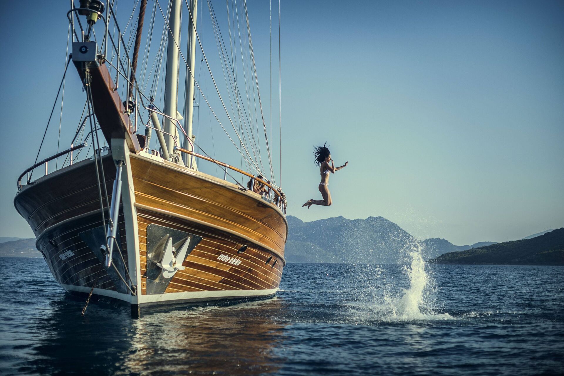 A person in swimwear jumps off the side of a wooden sailboat into calm blue water, creating a splash, with distant mountains and a clear sky in the background.