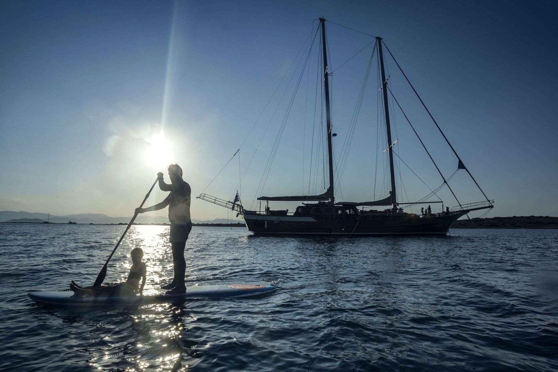 A person stands paddling on a board with a child sitting in front, silhouetted against the sun near a large sailboat anchored on calm waters, with distant mountains in the background.