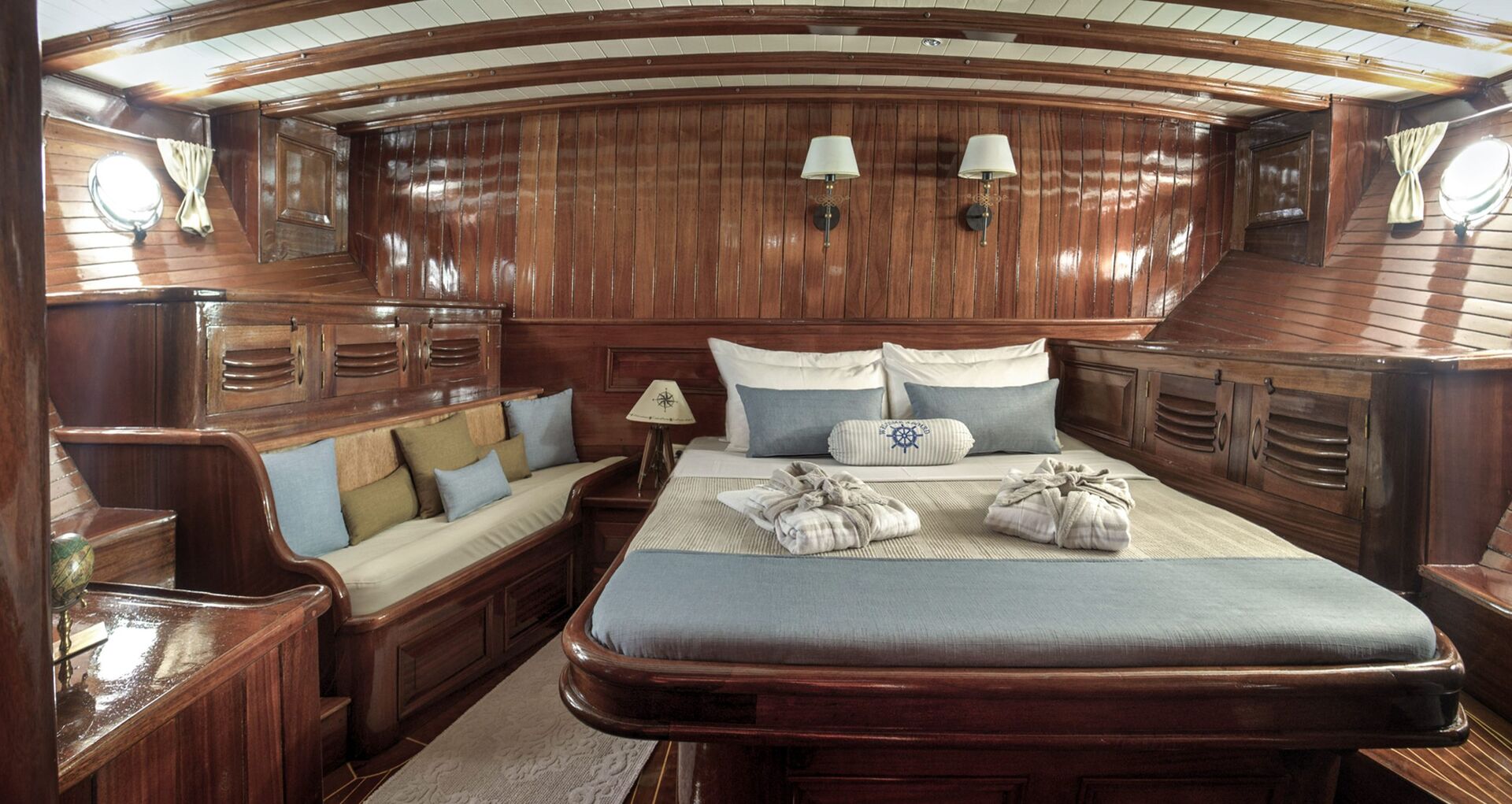 Wood-paneled yacht bedroom with a double bed, pillows, two towels artfully arranged on the bed, a cushioned bench with pillows on the left, wall lamps, small windows, and a cozy nautical ambiance.