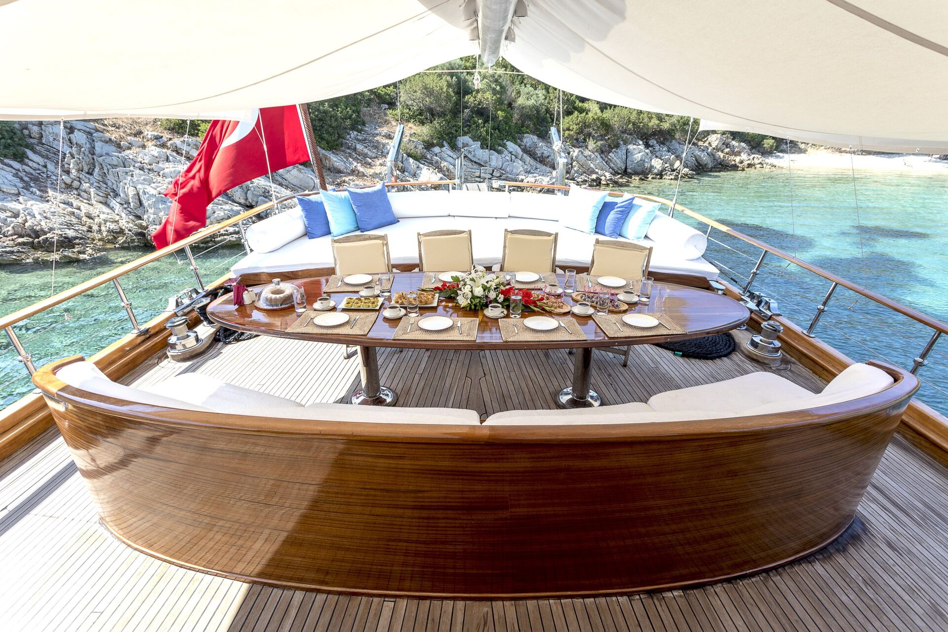 A luxurious yacht patio set for a meal with a wooden oval table, white cushioned chairs, and a sofa with blue pillows, overlooking clear turquoise water and a rocky shoreline.