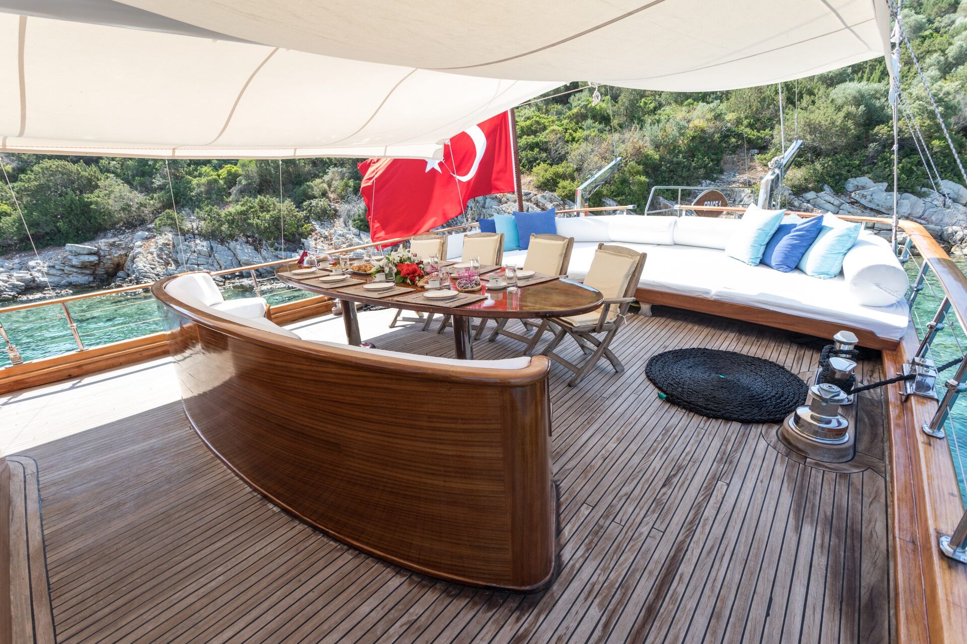 A yacht deck with a polished wooden table set for a meal, cream chairs, a curved wooden bench, white cushions, and a Turkish flag, surrounded by calm water and lush greenery.