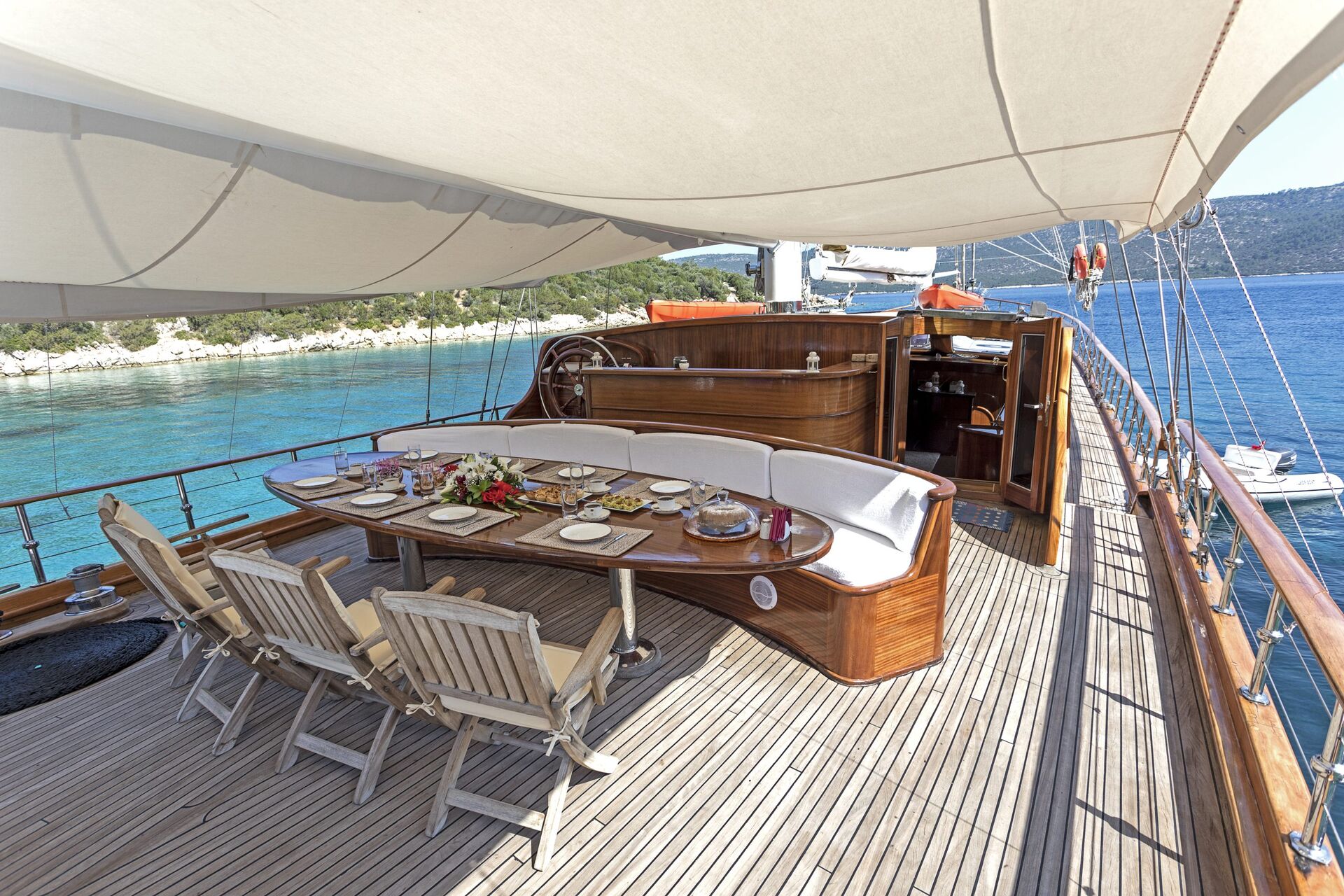 A luxurious yacht deck with a curved dining table set for a meal, cushioned seats, wooden chairs, and a sunshade overhead. Calm blue water and green hills are visible in the background.
