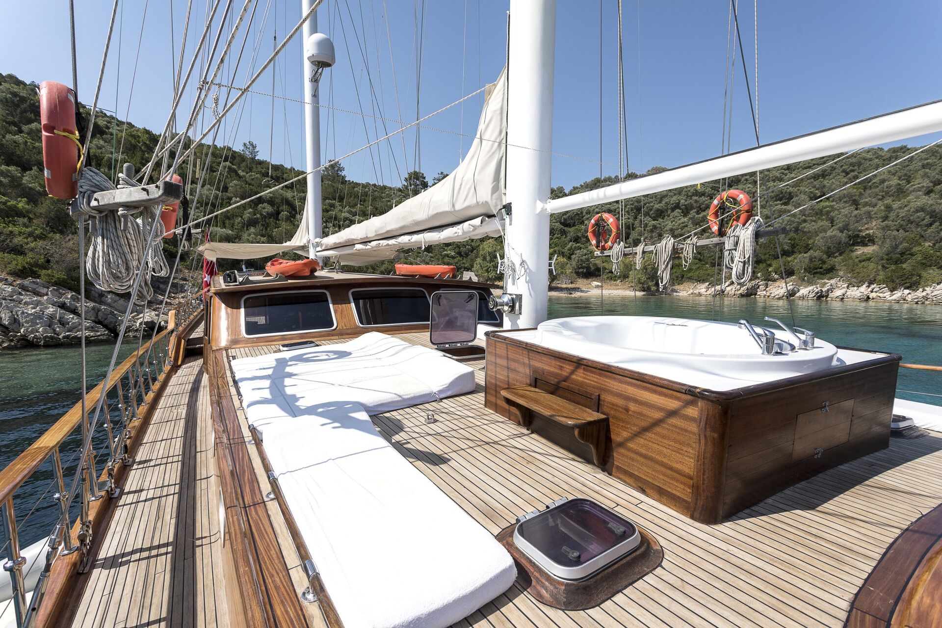 The deck of a wooden yacht features a sunbathing area with white cushions and a built-in jacuzzi, surrounded by clear blue water and lush green hills in the background under a sunny sky.