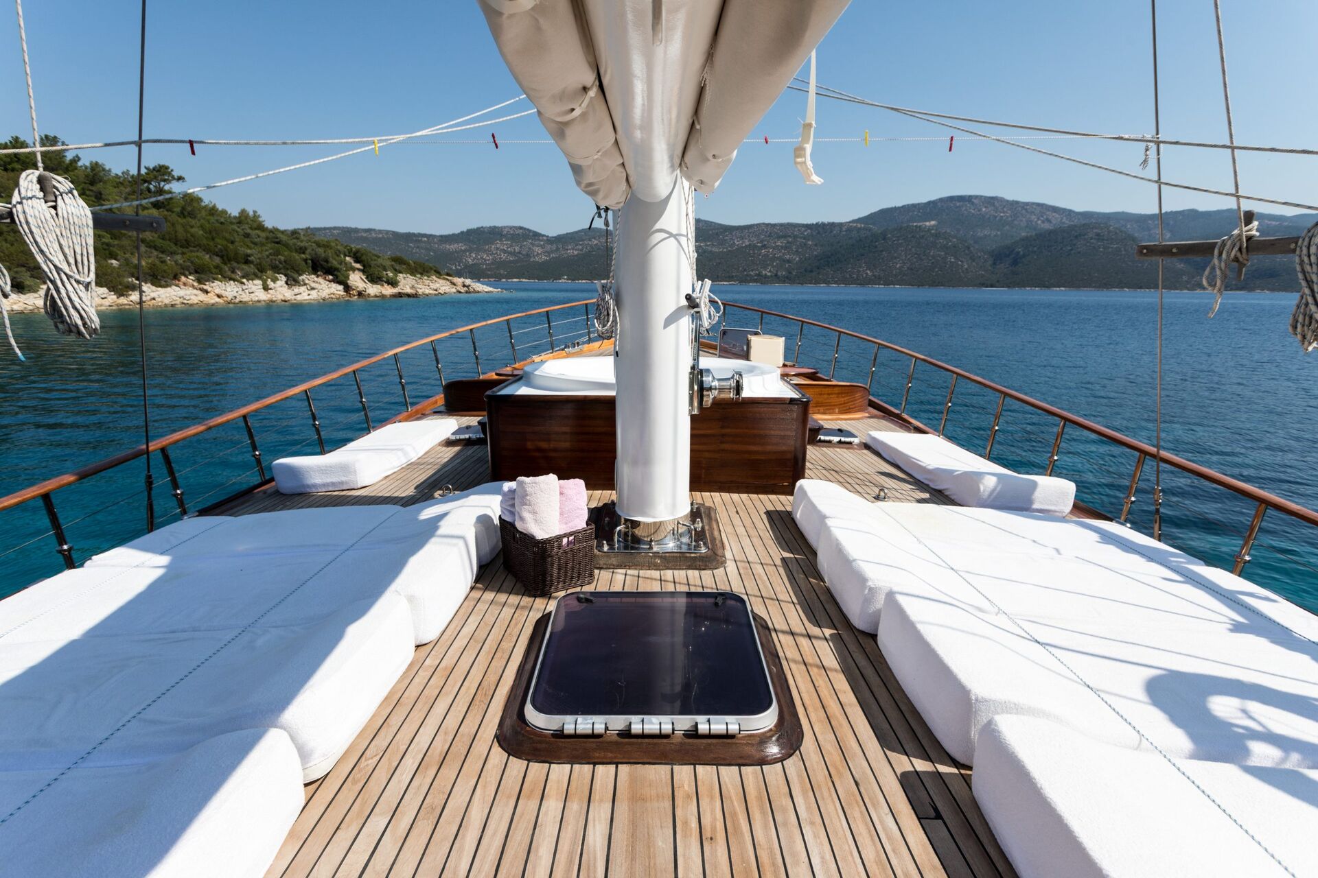 A spacious yacht deck with white cushioned lounge chairs and a view of calm blue water and distant mountains under a clear sky. Towels and a basket sit near the center mast.
