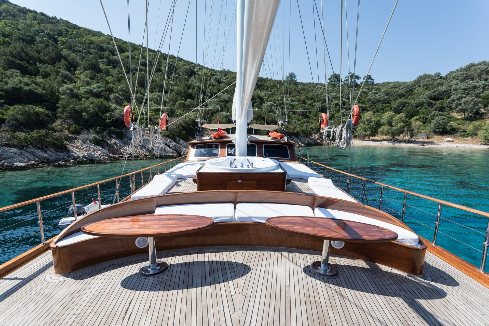View from the deck of a sailboat with curved wooden benches and white cushions, anchored near a lush, green shoreline and clear turquoise water under a sunny sky.