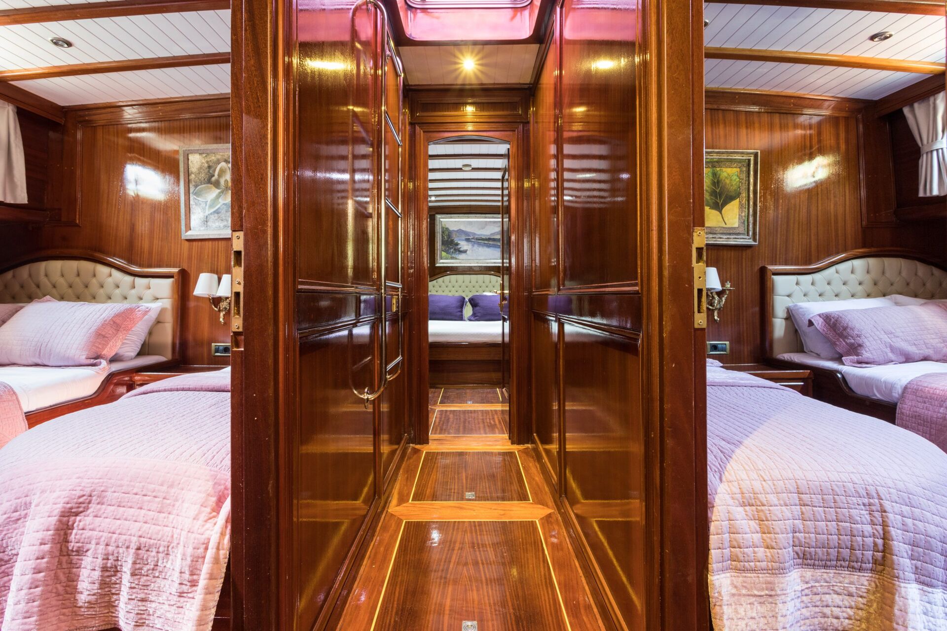 A hallway with polished wooden walls leads to two cozy bedrooms, each with twin beds covered in pale pink quilts and tufted headboards; paintings hang above the beds, and warm lighting fills the space.