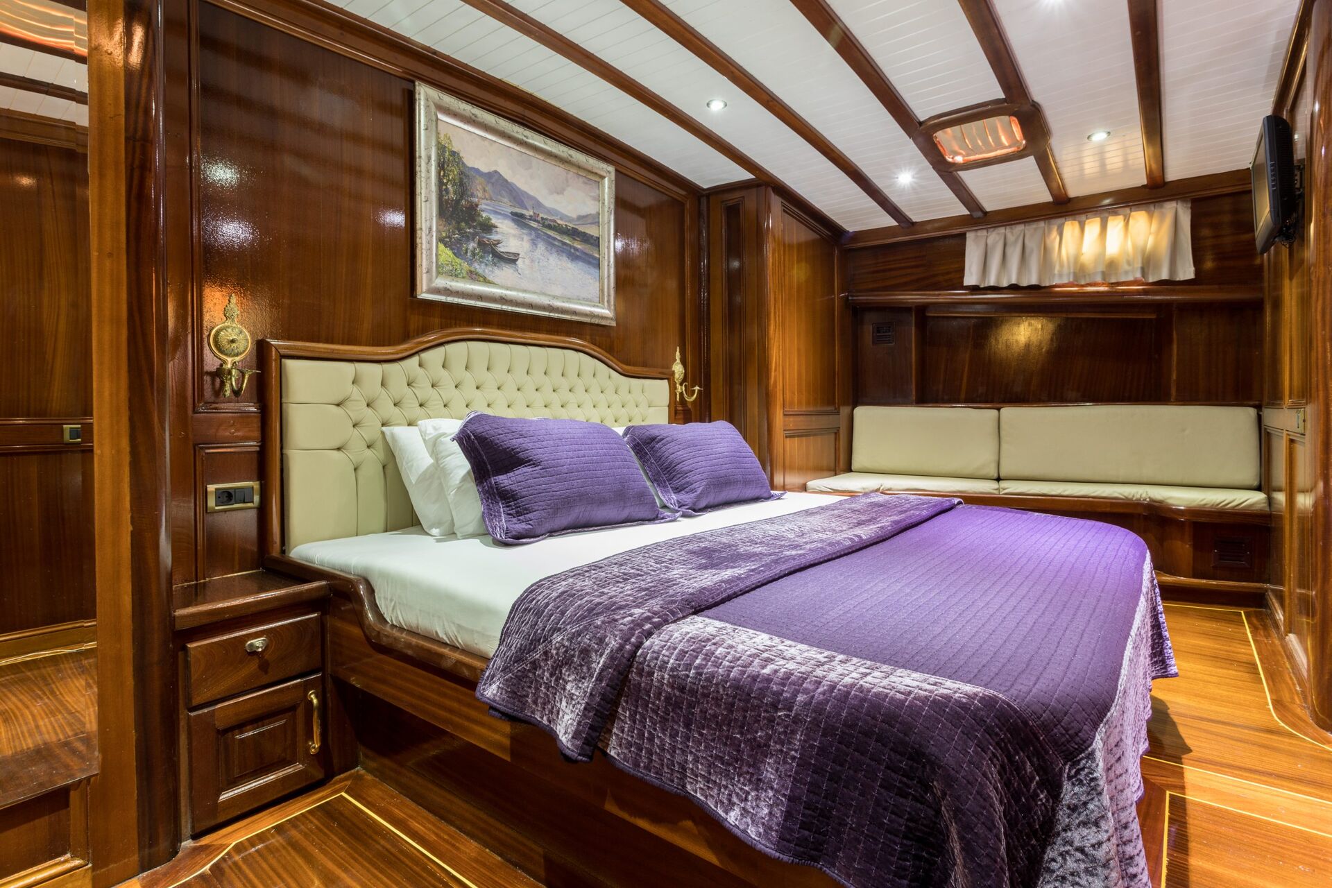 Luxurious bedroom with polished wood walls and ceiling, a large bed with a tufted headboard, purple bedding, a landscape painting above the bed, and a cushioned seating area along the wall. Warm lighting accents the space.