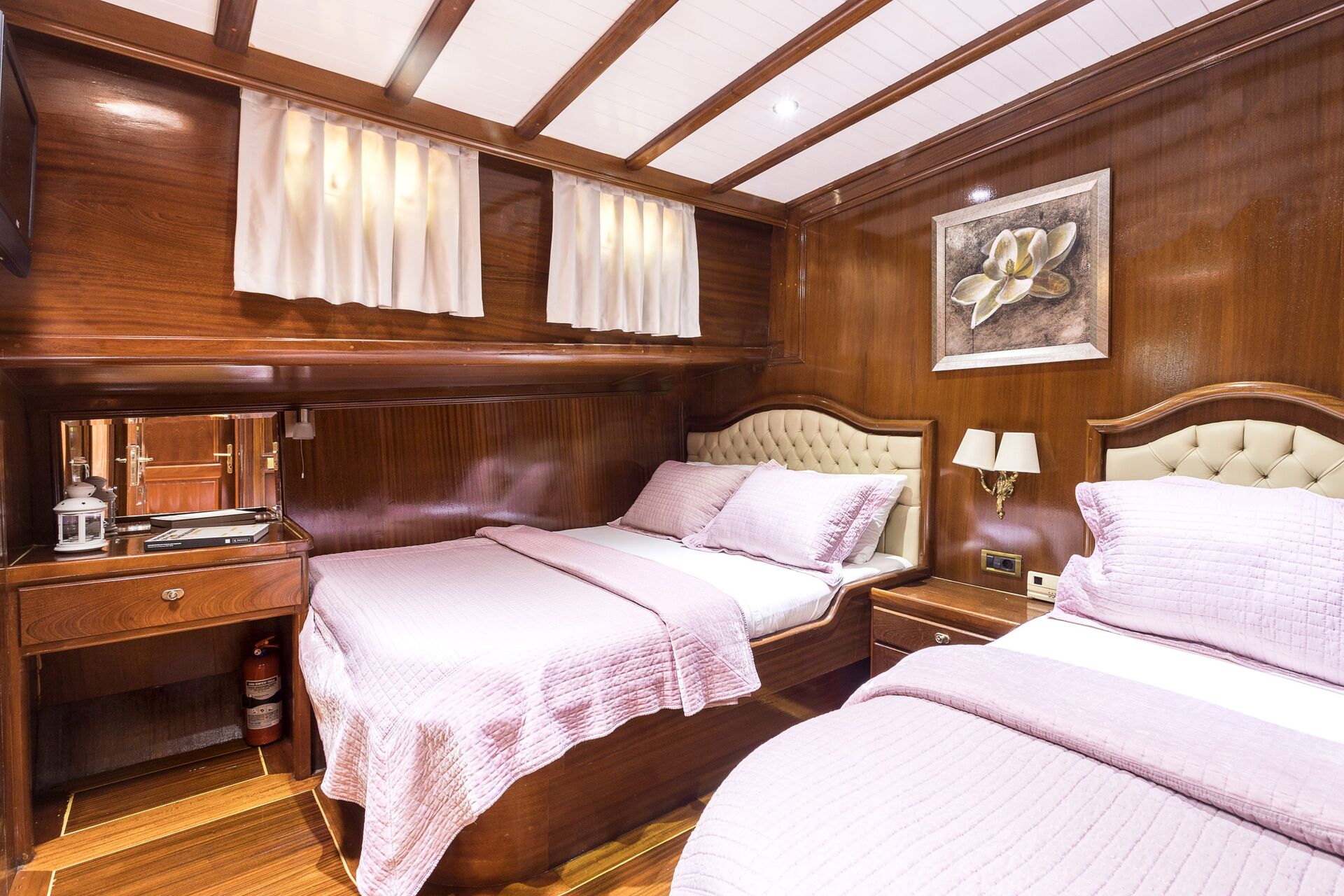 A cozy cabin bedroom with polished wood walls, two beds with tufted headboards and light purple bedding, a small wooden desk with a mirror, a lamp, and a flower painting on the wall.